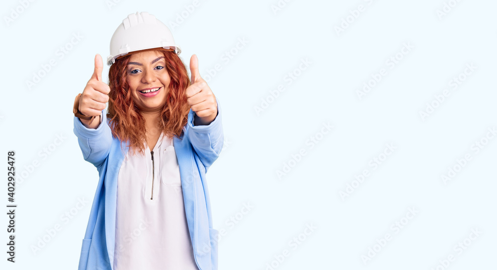 Young latin woman wearing architect hardhat approving doing positive gesture with hand, thumbs up smiling and happy for success. winner gesture. Young latin woman wearing architect hardhat approving doing positive gesture with hand, thumbs up smiling and happy for success. winner gesture.
