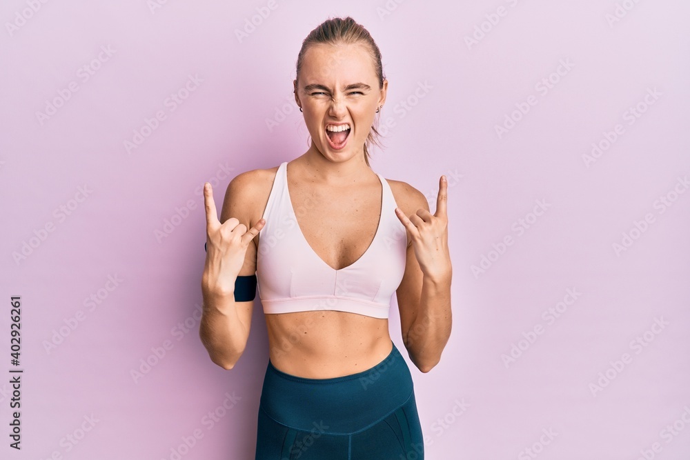 Beautiful blonde woman wearing sportswear and arm band shouting with crazy expression doing rock symbol with hands up. music star. heavy concept.