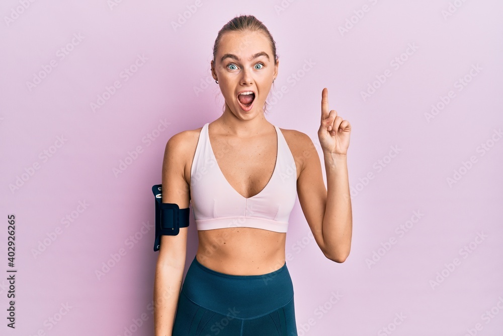 Beautiful blonde woman wearing sportswear and arm band pointing finger up with successful idea. exited and happy. number one.