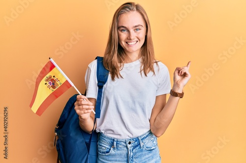 Beautiful blonde woman exchange student holding spanish flag smiling happy pointing with hand and finger to the side