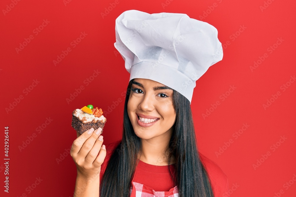 Young beautiful hispanic girl wearing chef hat holding homemade pastry looking positive and happy standing and smiling with a confident smile showing teeth