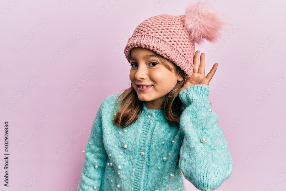 Little beautiful girl wearing wool sweater and cute winter hat smiling with hand over ear listening and hearing to rumor or gossip. deafness concept. Little beautiful girl wearing wool sweater and cute winter hat smiling with hand over ear listening and hearing to rumor or gossip. deafness concept.