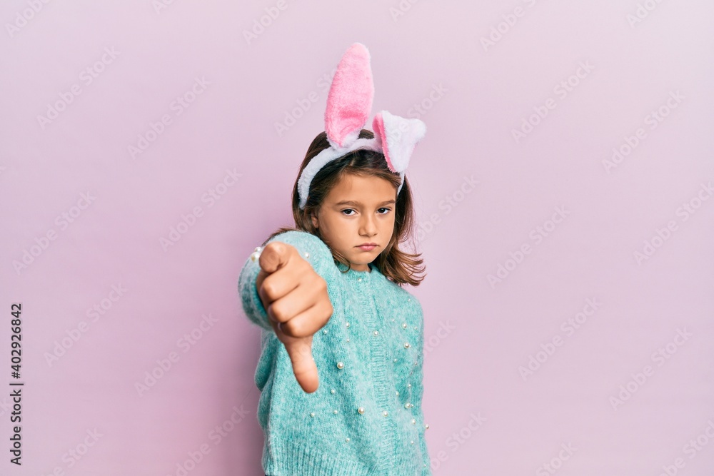 Little beautiful girl wearing cute easter bunny ears looking unhappy and angry showing rejection and negative with thumbs down gesture. bad expression. Little beautiful girl wearing cute easter bunny ears looking unhappy and angry showing rejection and negative with thumbs down gesture. bad expression.