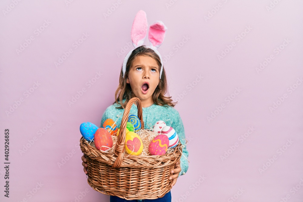 Little beautiful girl wearing cute easter bunny ears holding wicker basket with colored eggs angry and mad screaming frustrated and furious, shouting with anger looking up.