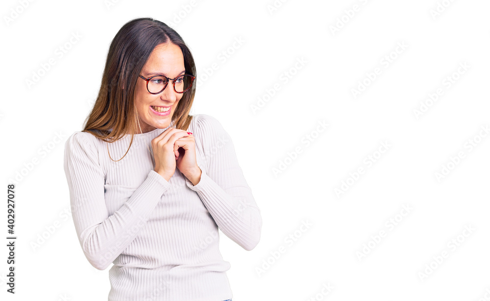 Beautiful brunette young woman wearing casual white sweater and glasses laughing nervous and excited with hands on chin looking to the side Beautiful brunette young woman wearing casual white sweater and glasses laughing nervous and excited with hands on chin looking to the side