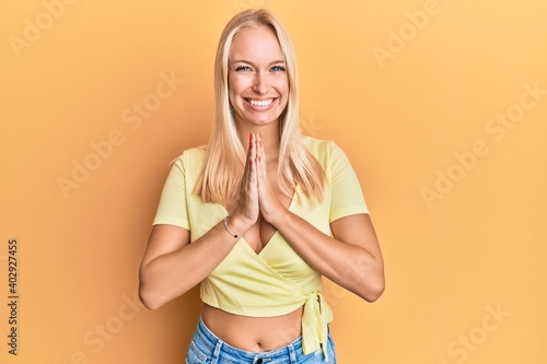 Young blonde girl wearing casual clothes praying with hands together asking for forgiveness smiling confident.