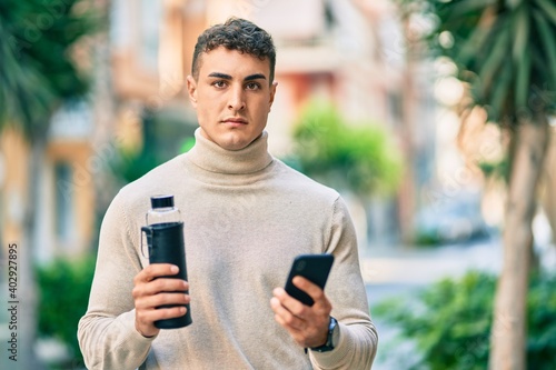 Young hispanic man with serious expression using smartphone and holding bottle of water at the city.