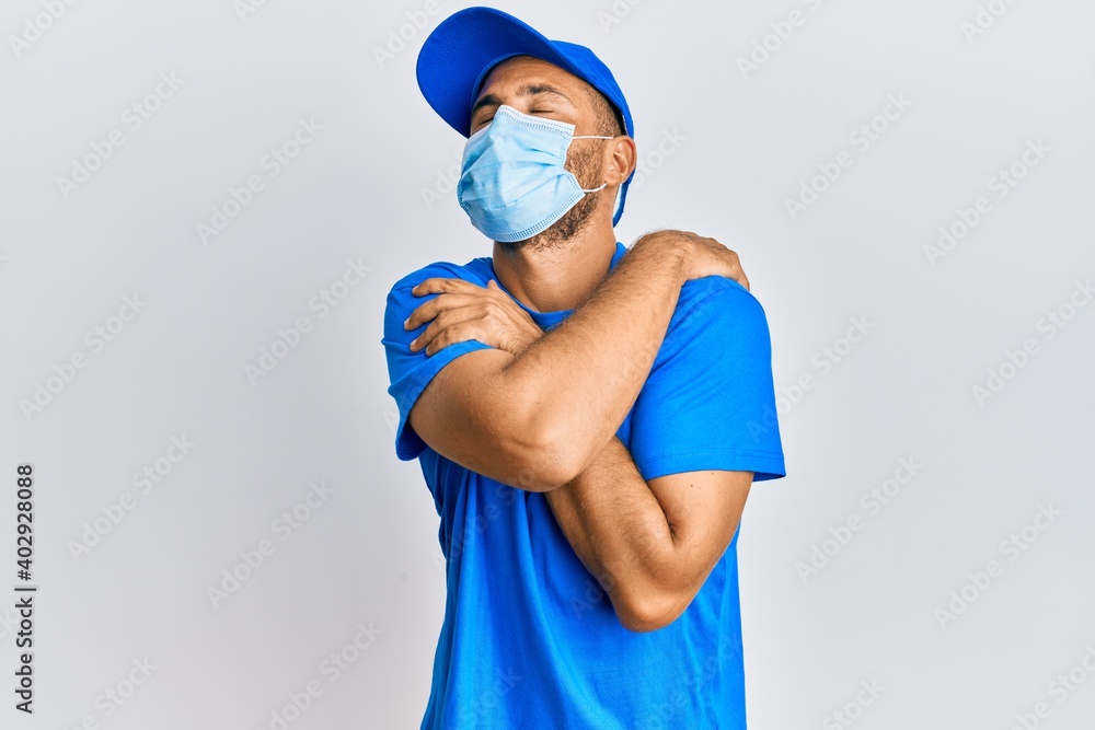 Handsome man with beard wearing courier uniform and medical mask hugging oneself happy and positive, smiling confident. self love and self care Handsome man with beard wearing courier uniform and medical mask hugging oneself happy and positive, smiling confident. self love and self care