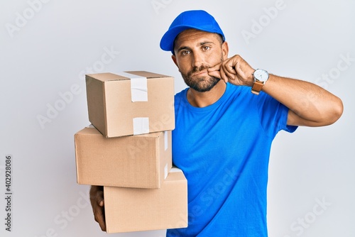 Handsome man with beard wearing courier uniform holding delivery packages mouth and lips shut as zip with fingers. secret and silent, taboo talking © Krakenimages.com