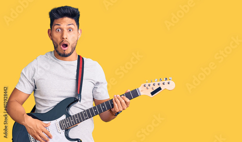 Handsome latin american young man playing electric guitar scared and amazed with open mouth for surprise, disbelief face