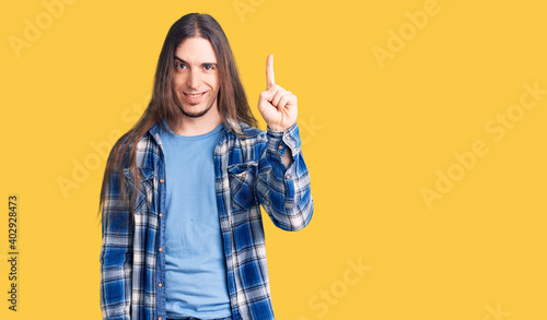 Young adult man with long hair wearing casual shirt showing and pointing up with finger number one while smiling confident and happy.