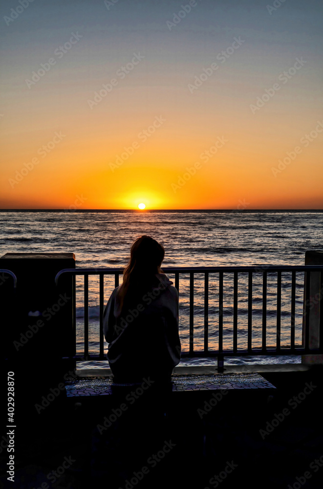 Fototapeta premium Woman sitting on bench with view of ocean and orange sun in San Diego California