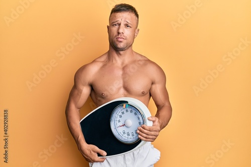 Handsome muscle man holding weight machine to balance weight loss clueless and confused expression. doubt concept.