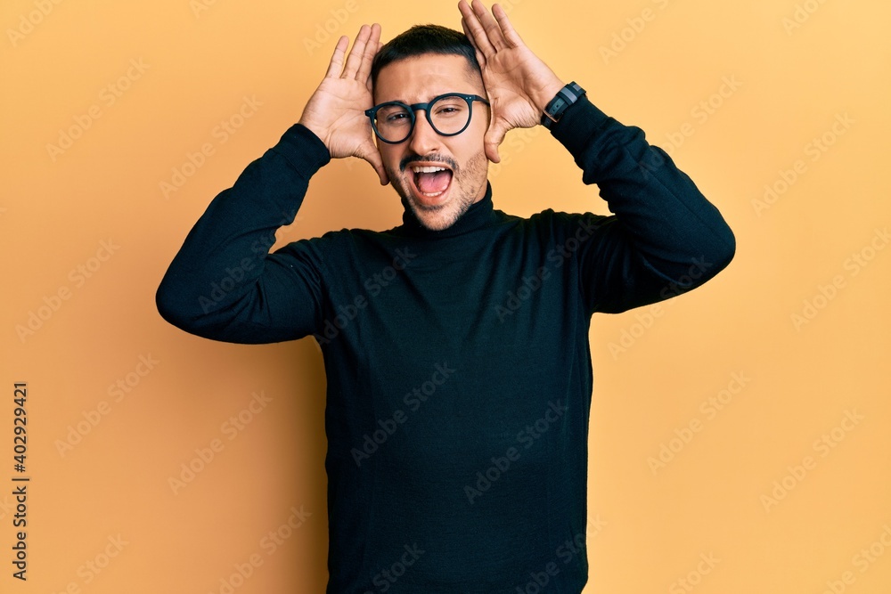 Handsome man with tattoos wearing turtleneck sweater and glasses smiling cheerful playing peek a boo with hands showing face. surprised and exited Handsome man with tattoos wearing turtleneck sweater and glasses smiling cheerful playing peek a boo with hands showing face. surprised and exited