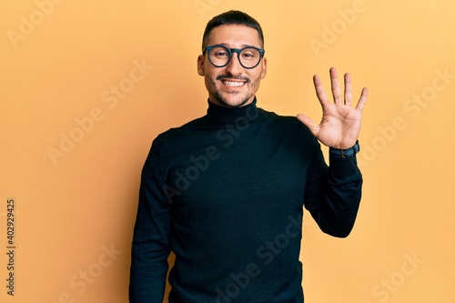 Handsome man with tattoos wearing turtleneck sweater and glasses showing and pointing up with fingers number five while smiling confident and happy.