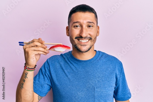 Handsome man with tattoos eating tuna sushi using chopsticks looking positive and happy standing and smiling with a confident smile showing teeth