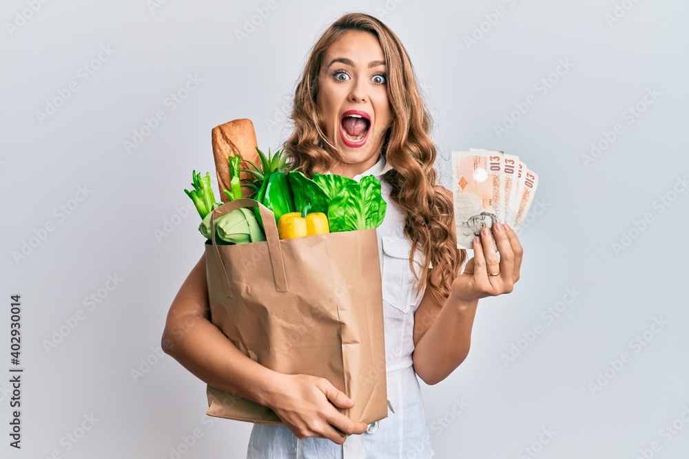 Young blonde girl holding paper bag with groceries and pounds celebrating crazy and amazed for success with open eyes screaming excited. Young blonde girl holding paper bag with groceries and pounds celebrating crazy and amazed for success with open eyes screaming excited.