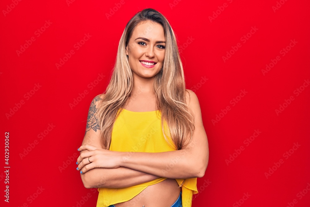 Young beautiful blonde woman wearing yellow t-shirt standing over isolated red background happy face smiling with crossed arms looking at the camera. Positive person. Young beautiful blonde woman wearing yellow t-shirt standing over isolated red background happy face smiling with crossed arms looking at the camera. Positive person.