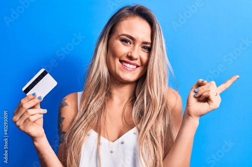 Young beautiful blonde woman holding credit card over isolated blue background smiling happy pointing with hand and finger to the side