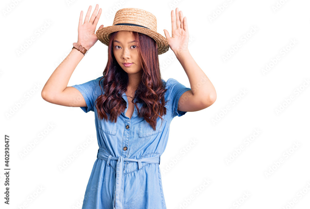 Young beautiful chinese girl wearing summer hat doing bunny ears gesture with hands palms looking cynical and skeptical. easter rabbit concept. Young beautiful chinese girl wearing summer hat doing bunny ears gesture with hands palms looking cynical and skeptical. easter rabbit concept.