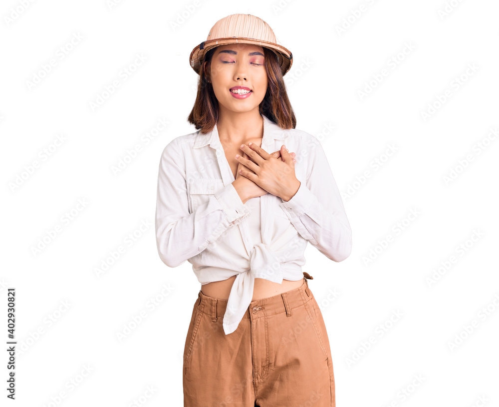 Young beautiful chinese girl wearing explorer hat smiling with hands on chest with closed eyes and grateful gesture on face. health concept. Young beautiful chinese girl wearing explorer hat smiling with hands on chest with closed eyes and grateful gesture on face. health concept.