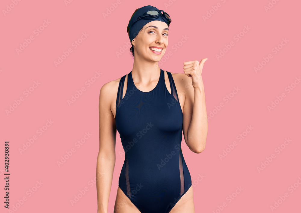 Young beautiful hispanic woman wearing swimwear and swimmer glasses smiling with happy face looking and pointing to the side with thumb up. Young beautiful hispanic woman wearing swimwear and swimmer glasses smiling with happy face looking and pointing to the side with thumb up.