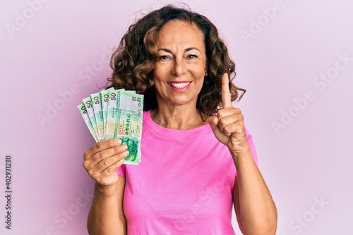 Middle age hispanic woman holding 50 hong kong dollars banknotes smiling with an idea or question pointing finger with happy face, number one