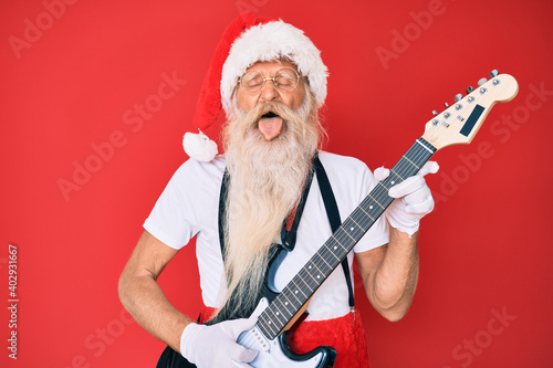 Old senior man wearing santa claus costume playing electric guitar sticking tongue out happy with funny expression.