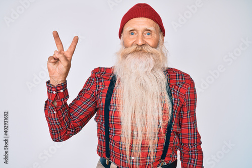 Old senior man with grey hair and long beard wearing hipster look with wool cap smiling looking to the camera showing fingers doing victory sign. number two.