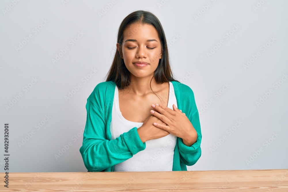 Beautiful hispanic woman wearing casual clothes sitting on the table smiling with hands on chest, eyes closed with grateful gesture on face. health concept. Beautiful hispanic woman wearing casual clothes sitting on the table smiling with hands on chest, eyes closed with grateful gesture on face. health concept.