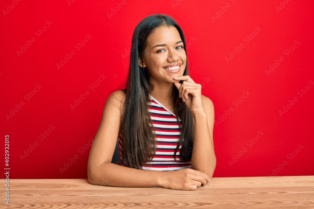 Beautiful hispanic woman wearing casual clothes sitting on the table looking confident at the camera with smile with crossed arms and hand raised on chin. thinking positive. Beautiful hispanic woman wearing casual clothes sitting on the table looking confident at the camera with smile with crossed arms and hand raised on chin. thinking positive.