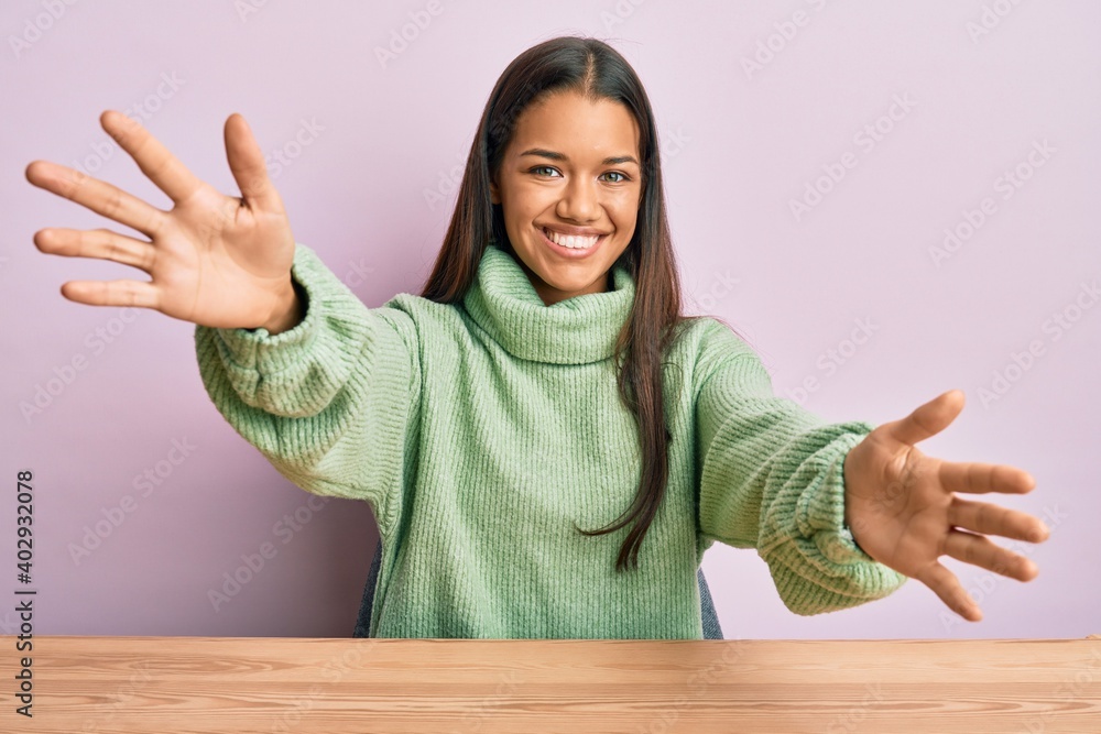 Beautiful hispanic woman wearing casual clothes sitting on the table looking at the camera smiling with open arms for hug. cheerful expression embracing happiness. Beautiful hispanic woman wearing casual clothes sitting on the table looking at the camera smiling with open arms for hug. cheerful expression embracing happiness.