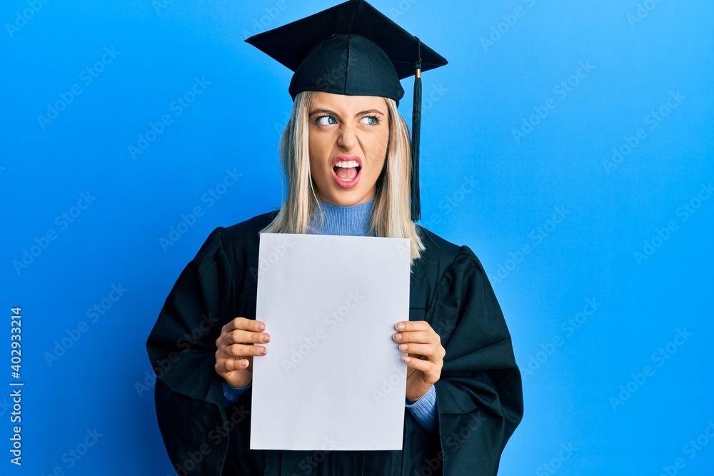 Beautiful blonde woman wearing graduation cap and ceremony robe holding blank banner angry and mad screaming frustrated and furious, shouting with anger. rage and aggressive concept. Beautiful blonde woman wearing graduation cap and ceremony robe holding blank banner angry and mad screaming frustrated and furious, shouting with anger. rage and aggressive concept.