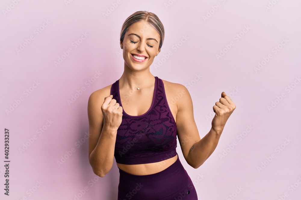 Beautiful blonde woman wearing sportswear over pink background excited for success with arms raised and eyes closed celebrating victory smiling. winner concept. Beautiful blonde woman wearing sportswear over pink background excited for success with arms raised and eyes closed celebrating victory smiling. winner concept.