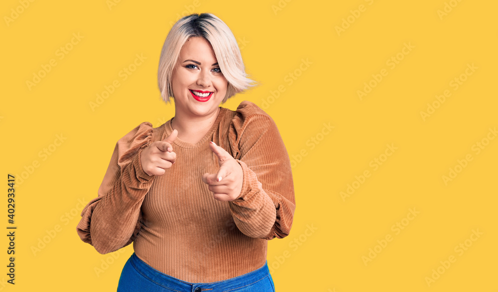 Young blonde plus size woman wearing casual sweater pointing fingers to camera with happy and funny face. good energy and vibes. Young blonde plus size woman wearing casual sweater pointing fingers to camera with happy and funny face. good energy and vibes.