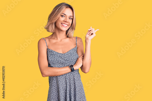 Beautiful caucasian woman wearing summer dress with a big smile on face, pointing with hand and finger to the side looking at the camera.