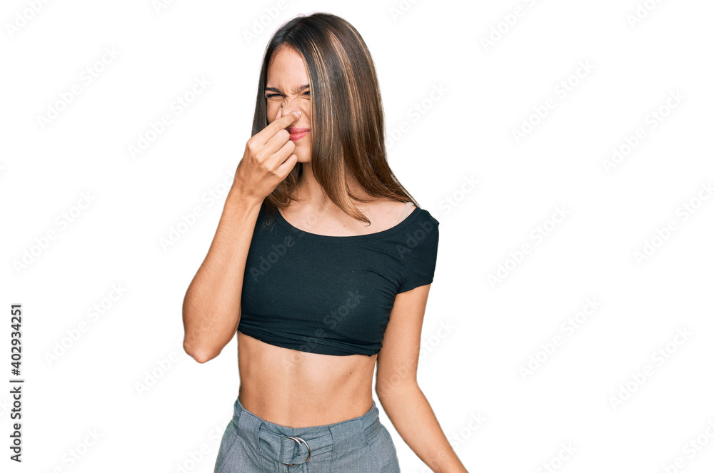 Young brunette woman wearing casual clothes smelling something stinky and disgusting, intolerable smell, holding breath with fingers on nose. bad smell