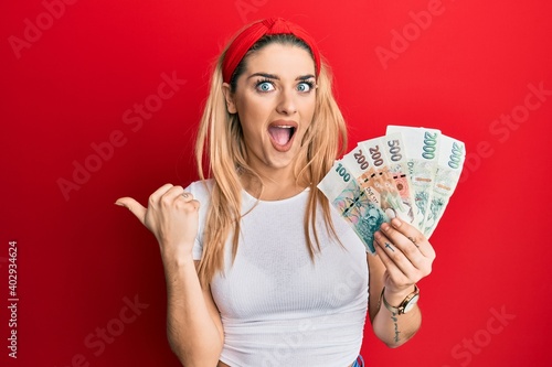 Young caucasian woman holding czech koruna banknotes pointing thumb up to the side smiling happy with open mouth