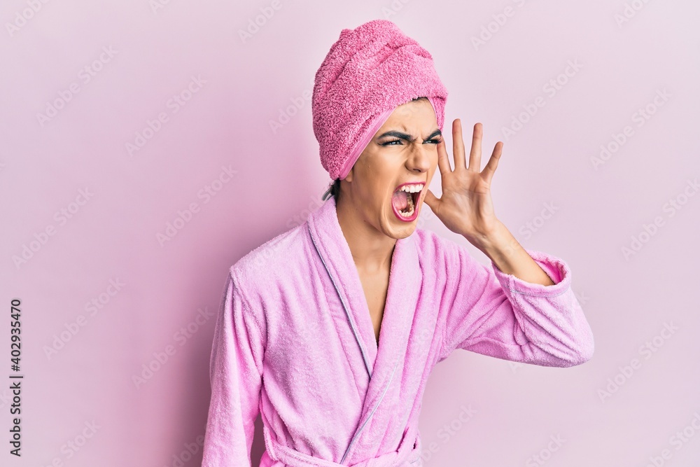 Young man wearing woman make up wearing shower towel on head and bathrobe shouting and screaming loud to side with hand on mouth. communication concept. Young man wearing woman make up wearing shower towel on head and bathrobe shouting and screaming loud to side with hand on mouth. communication concept.