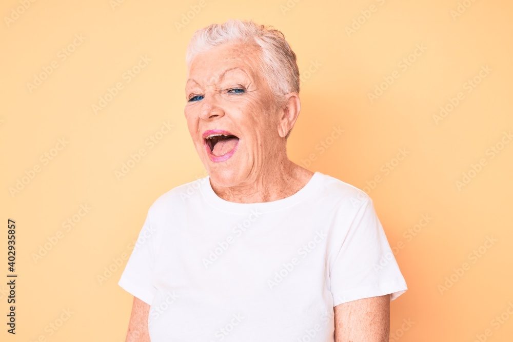 Senior beautiful woman with blue eyes and grey hair wearing classic white tshirt over yellow background winking looking at the camera with sexy expression, cheerful and happy face. Senior beautiful woman with blue eyes and grey hair wearing classic white tshirt over yellow background winking looking at the camera with sexy expression, cheerful and happy face.