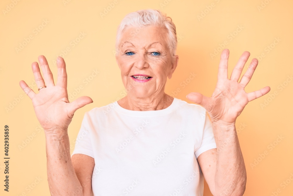 Senior beautiful woman with blue eyes and grey hair wearing classic white tshirt over yellow background showing and pointing up with fingers number ten while smiling confident and happy. Senior beautiful woman with blue eyes and grey hair wearing classic white tshirt over yellow background showing and pointing up with fingers number ten while smiling confident and happy.