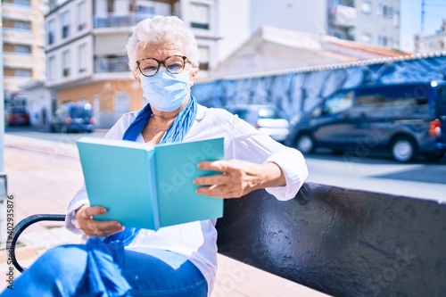 Elder senior woman with grey hair wearing coronavirus safety mask outdoors sitting on a bench reading a book