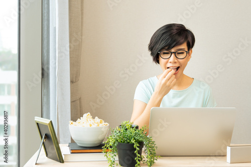 Beautiful and stylish young asian woman enjoy having popcorn snack while using computer laptop to working from home during the pandemic lockdown. Covid 19 New normal, Stay home, Stay healthy concept.