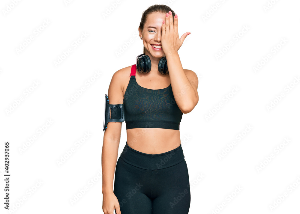 Beautiful blonde woman wearing gym clothes and using headphones covering one eye with hand, confident smile on face and surprise emotion.