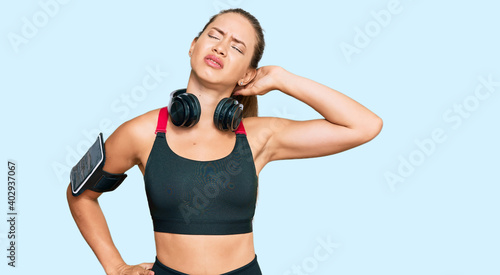 Beautiful blonde woman wearing gym clothes and using headphones suffering of neck ache injury, touching neck with hand, muscular pain