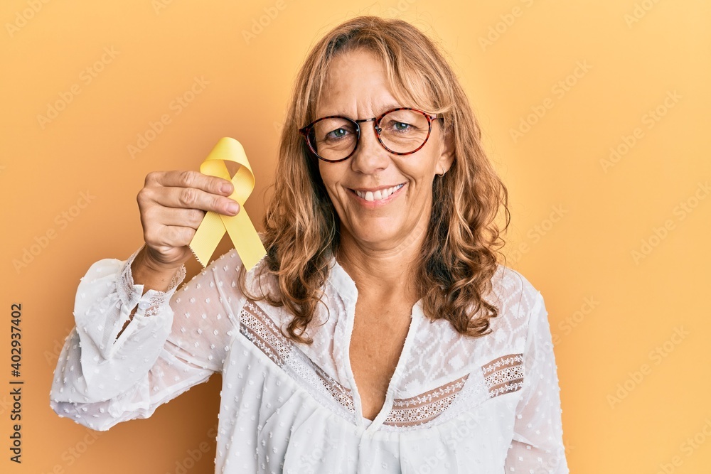 Middle age blonde woman holding suicide prevention yellow ribbon looking positive and happy standing and smiling with a confident smile showing teeth Middle age blonde woman holding suicide prevention yellow ribbon looking positive and happy standing and smiling with a confident smile showing teeth