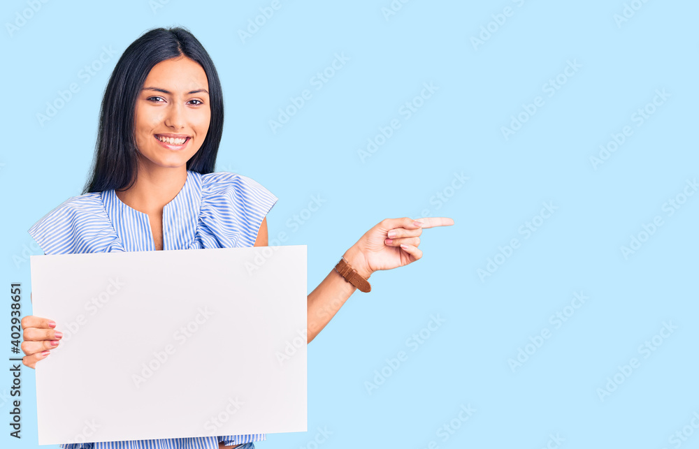 Young beautiful latin girl holding blank empty banner smiling happy pointing with hand and finger to the side Young beautiful latin girl holding blank empty banner smiling happy pointing with hand and finger to the side