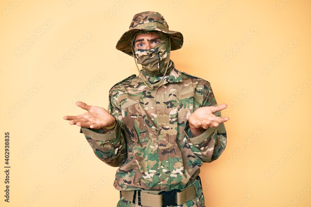Fototapeta premium Young handsome man wearing camouflage army uniform and balaclava clueless and confused expression with arms and hands raised. doubt concept.