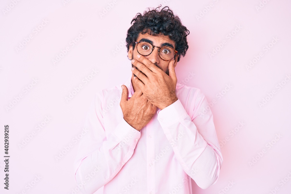 Handsome young man with curly hair and bear wearing business shirt and glasses shocked covering mouth with hands for mistake. secret concept. Handsome young man with curly hair and bear wearing business shirt and glasses shocked covering mouth with hands for mistake. secret concept.
