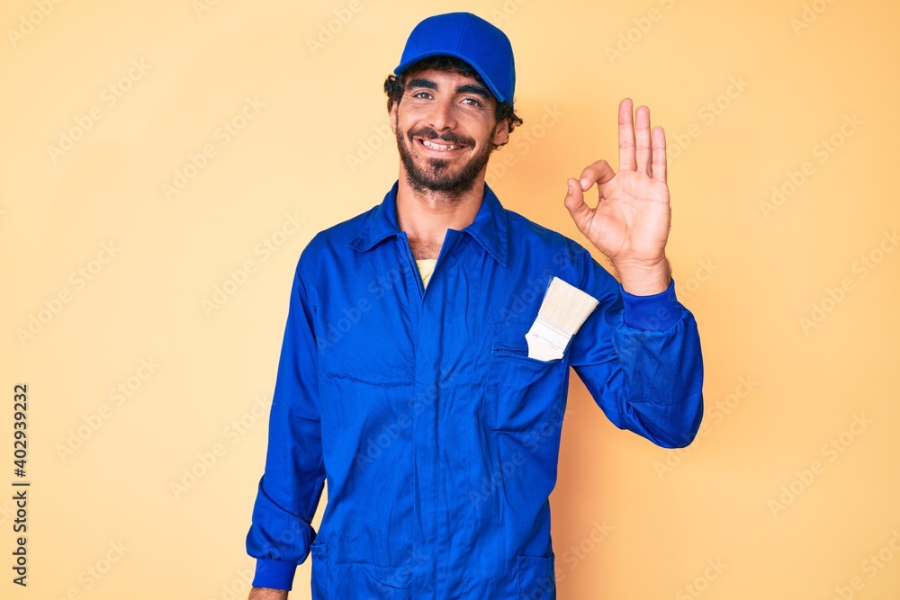 Handsome young man with curly hair and bear wearing builder jumpsuit uniform smiling positive doing ok sign with hand and fingers. successful expression.